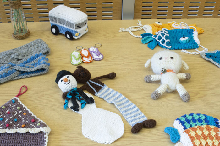 A wooden table with various knitted and crocheted items displayed on it, including snowman, bear, sheep, and VW campervan plushies, flip flop design keychains, headbands, and wall hangings.