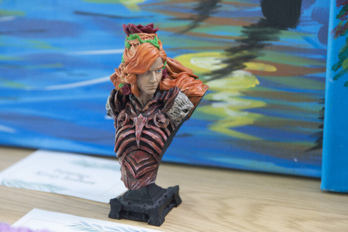 Image of a small model bust of a male figure with long ginger hair with flowers and plants running through it. The figure is wearing plate armour.