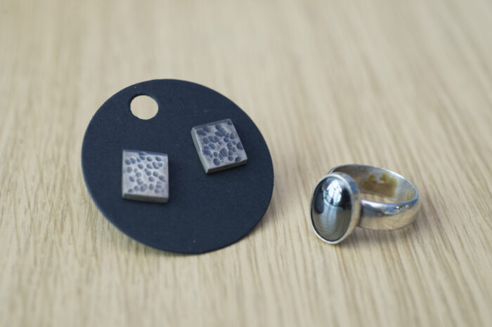 Two pieces of jewellery places on a wooden table. There are square earrings with a speckled blue design on a round stand, and a sliver ring with a dark inset gemstone.