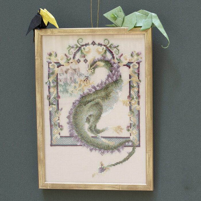 A framed rectangular cross stitch of a green dragon with flames coming out of its mouth. The dragon is framed within a stitched border of purple, yellow, blue and green design. An origami toucan and chameleon are placed on top of the framed tapestry.