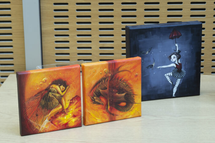 Two smaller square canvases painted in oranges, reds and yellows. One depicts a curled up fairy and the other is a curled up dragon. There is a third canvas which is square and slightly larger. It depicts a female figure dancing and holding a red umbrella with two birds near her.