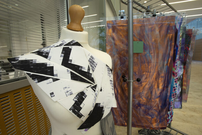 A mannequin stand with a black and white garment on display in the foreground. Behind it, textiles samples are visible on a railing.