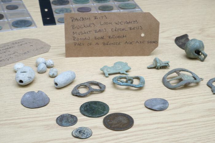 Various items found through metal detecting are displayed on a table. They include coins, brooches, buckles and loom weights.