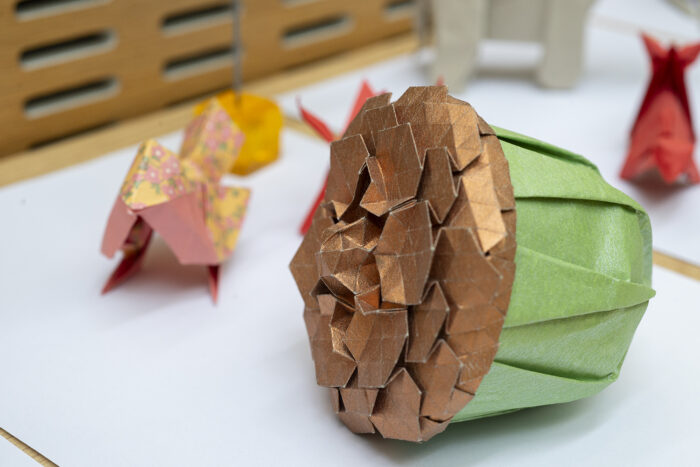 Origami Acorn in green and bronze paper in the foreground, with other origami creations out of focus in the background.